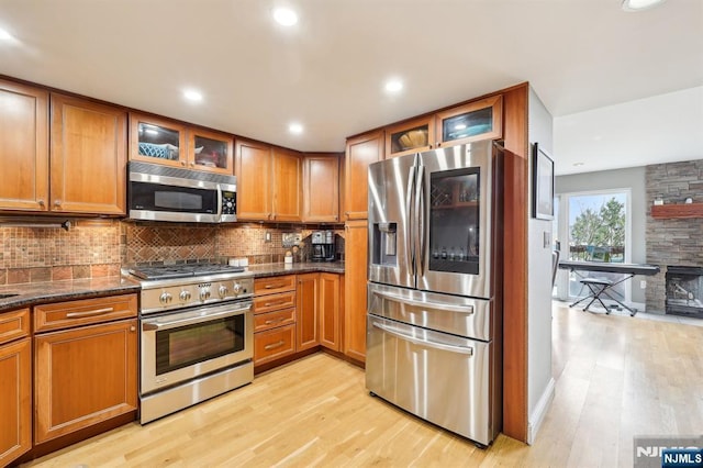 kitchen with decorative backsplash, glass insert cabinets, brown cabinets, light wood finished floors, and appliances with stainless steel finishes