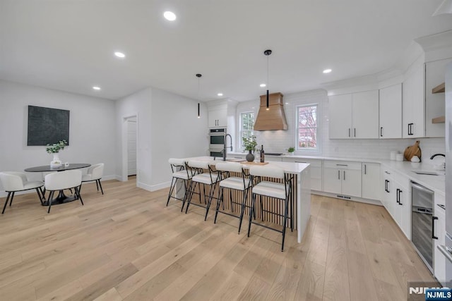 kitchen with light countertops, premium range hood, a center island with sink, decorative backsplash, and light wood-style flooring