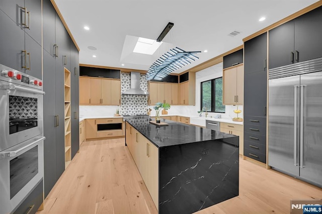kitchen with a skylight, wall chimney exhaust hood, light wood finished floors, and appliances with stainless steel finishes