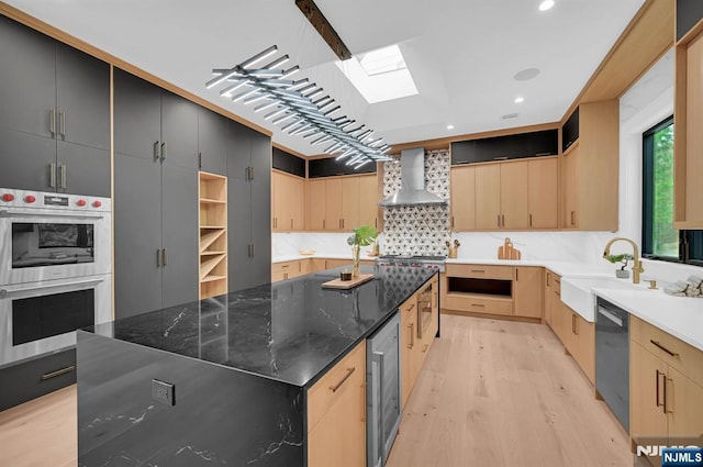 kitchen with double oven, beverage cooler, light brown cabinets, a sink, and wall chimney exhaust hood