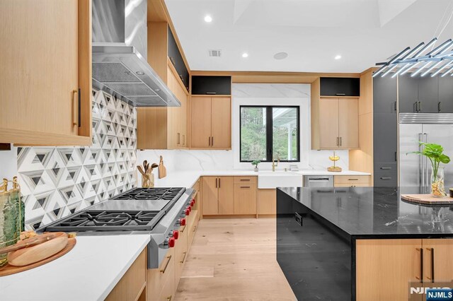 kitchen featuring light brown cabinetry, a sink, wall chimney exhaust hood, decorative backsplash, and appliances with stainless steel finishes