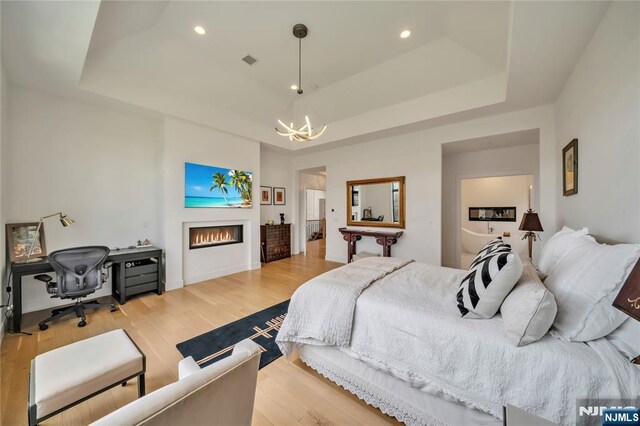 bedroom with a chandelier, light wood-type flooring, a warm lit fireplace, and a tray ceiling