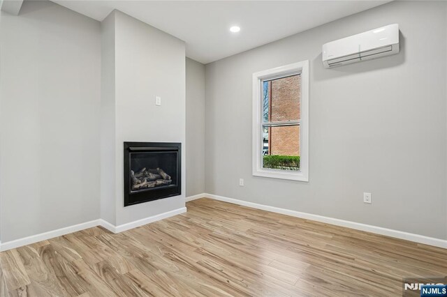 unfurnished living room featuring a wall unit AC, light wood-type flooring, a glass covered fireplace, baseboards, and recessed lighting