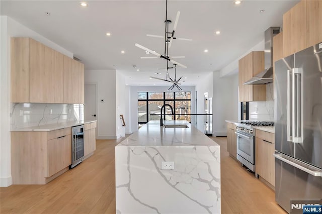 kitchen featuring wall chimney range hood, a sink, high end appliances, light brown cabinetry, and modern cabinets