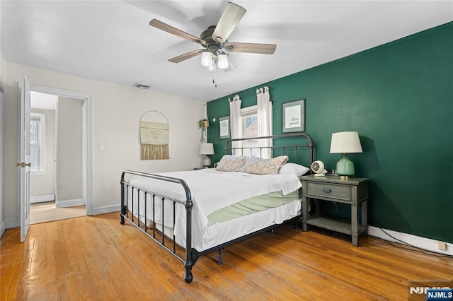 bedroom with a ceiling fan, wood finished floors, a baseboard heating unit, baseboards, and visible vents