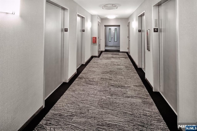 hall featuring baseboards, dark colored carpet, elevator, and a textured wall