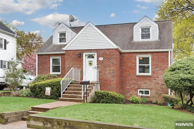 cape cod-style house featuring brick siding, a chimney, a front yard, and roof with shingles