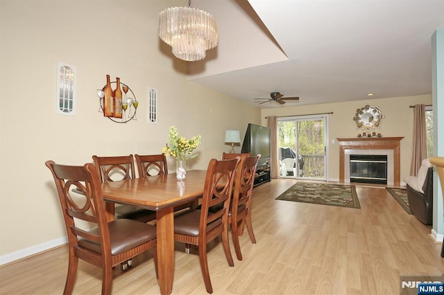 dining space with baseboards, light wood-type flooring, ceiling fan with notable chandelier, and a glass covered fireplace