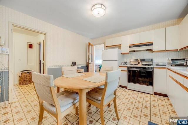 kitchen with white appliances, wallpapered walls, under cabinet range hood, light countertops, and white cabinets