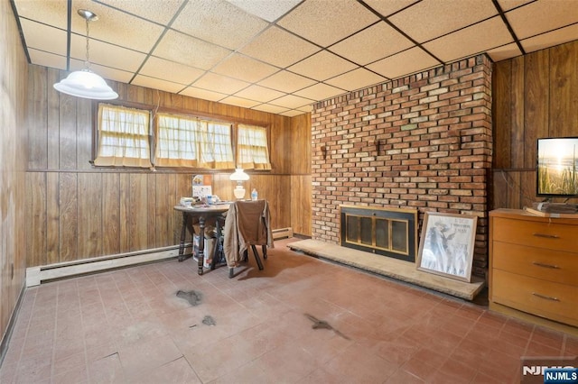 interior space with wooden walls, baseboard heating, a drop ceiling, and a brick fireplace
