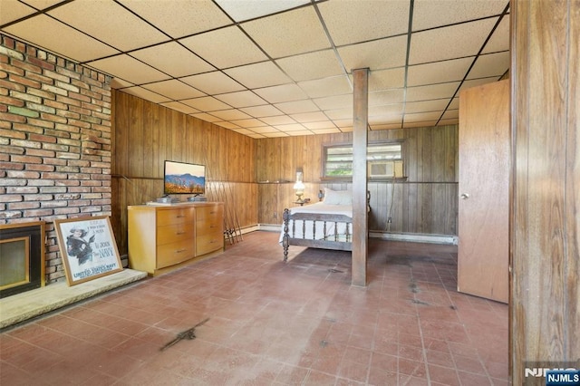 bedroom featuring wooden walls, baseboards, a brick fireplace, and a paneled ceiling