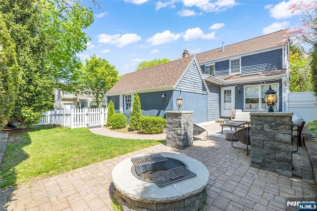 back of property with a fire pit, fence, roof with shingles, a patio area, and a lawn