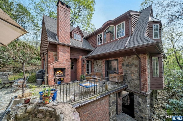 back of house with a shingled roof, a fireplace, stone siding, a chimney, and a patio