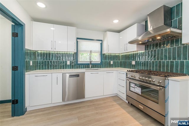 kitchen featuring light wood-type flooring, wall chimney exhaust hood, white cabinetry, a sink, and stainless steel appliances