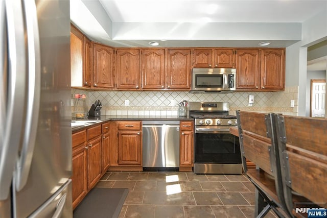 kitchen featuring backsplash, stainless steel appliances, dark countertops, and brown cabinetry