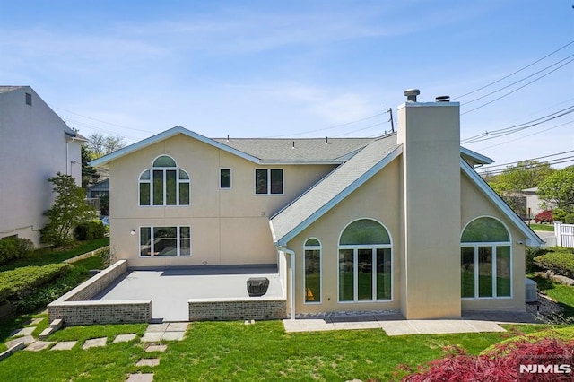 back of house with a chimney, a lawn, a patio area, a shingled roof, and stucco siding