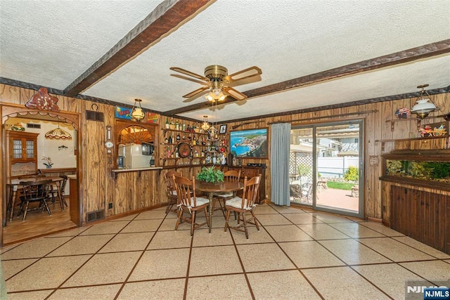 dining space featuring wooden walls, a ceiling fan, a textured ceiling, and beamed ceiling