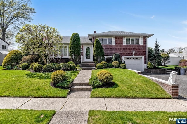 split level home featuring a front lawn, a chimney, brick siding, an attached garage, and driveway