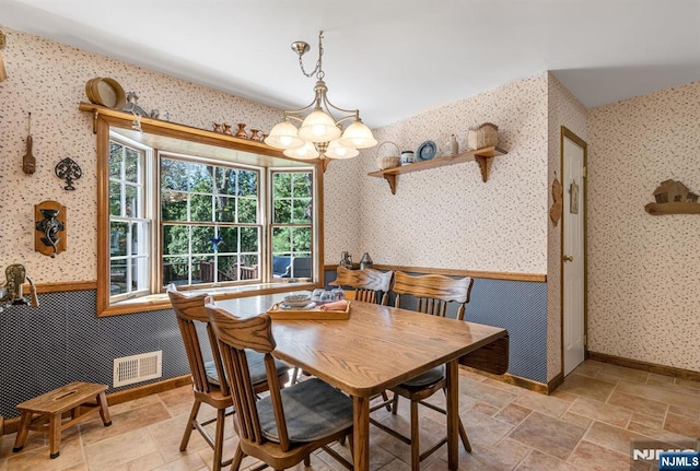 dining space with an inviting chandelier, wainscoting, wallpapered walls, stone finish flooring, and visible vents