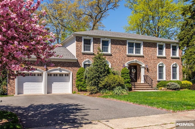 colonial house featuring a garage, aphalt driveway, a front yard, brick siding, and a shingled roof