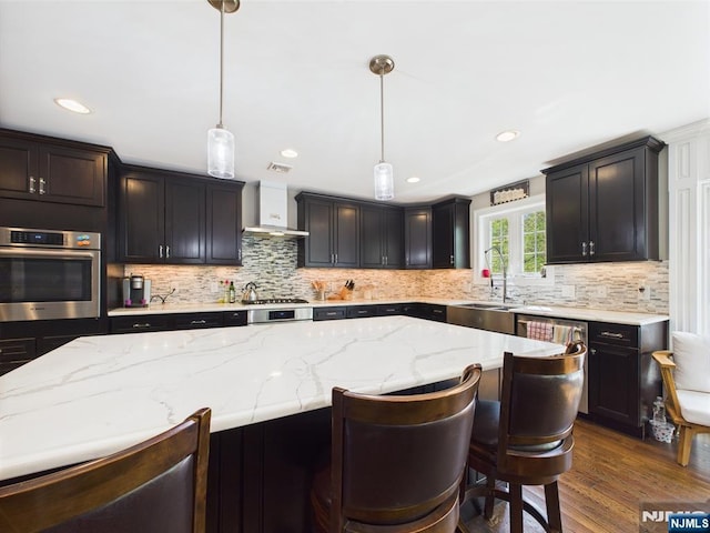 kitchen with wall chimney range hood, light stone counters, a sink, stainless steel oven, and dark wood-type flooring