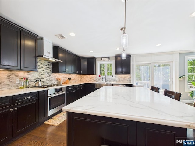 kitchen featuring wall chimney range hood, decorative backsplash, a kitchen island, a sink, and stainless steel oven