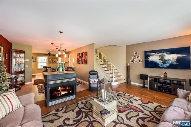 living room featuring ceiling fan, wood finished floors, stairway, baseboards, and a glass covered fireplace