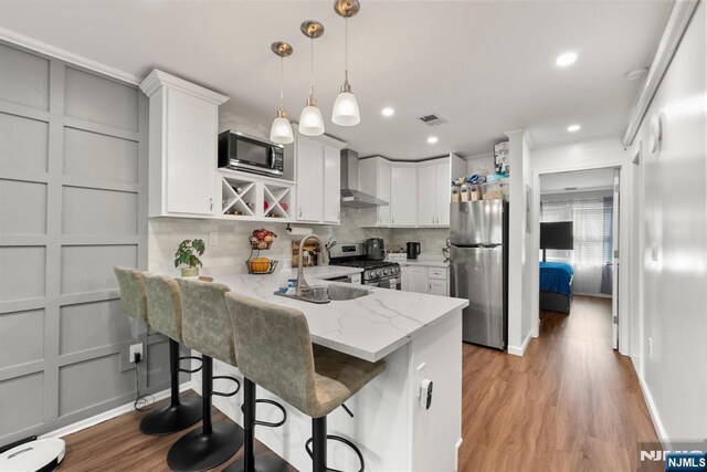 kitchen featuring visible vents, a peninsula, wall chimney range hood, a sink, and appliances with stainless steel finishes