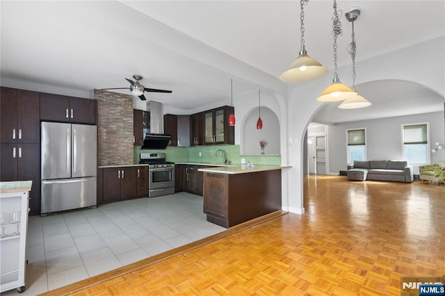 kitchen featuring dark brown cabinets, wall chimney exhaust hood, arched walkways, a ceiling fan, and appliances with stainless steel finishes