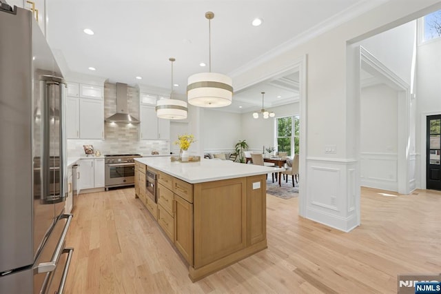 kitchen with appliances with stainless steel finishes, light wood finished floors, wall chimney exhaust hood, a kitchen island, and ornamental molding