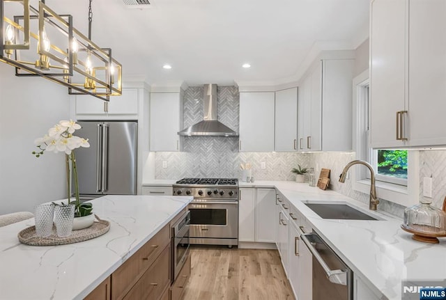 kitchen featuring high end appliances, wall chimney exhaust hood, white cabinets, a sink, and light wood-style flooring