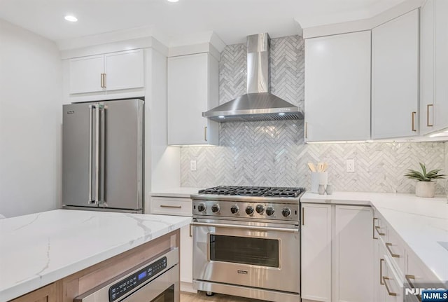 kitchen featuring wall chimney range hood, tasteful backsplash, light stone counters, stainless steel appliances, and white cabinetry