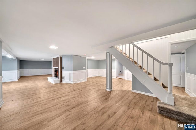 finished basement with stairs, wainscoting, a brick fireplace, a decorative wall, and light wood-type flooring