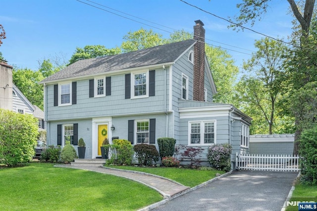 colonial-style house with a front lawn, an outdoor structure, roof with shingles, and a chimney