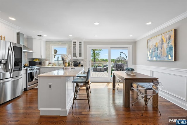 kitchen with wall chimney range hood, a breakfast bar, crown molding, glass insert cabinets, and appliances with stainless steel finishes