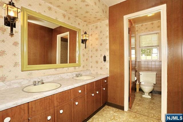full bath featuring a sink, double vanity, toilet, and wallpapered walls