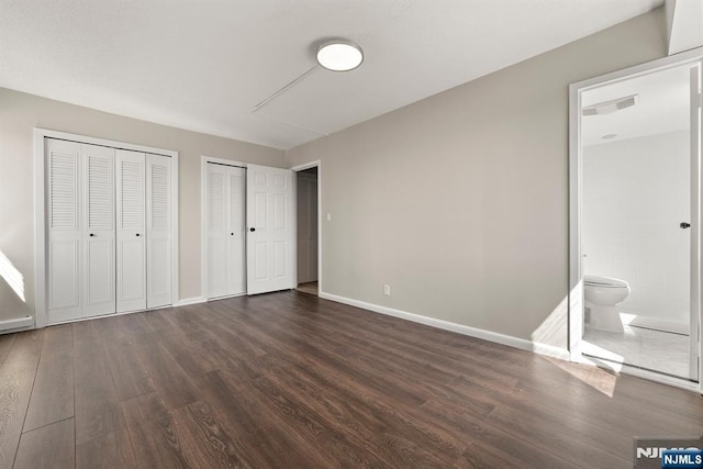 unfurnished bedroom with two closets, ensuite bathroom, baseboards, visible vents, and dark wood-style floors