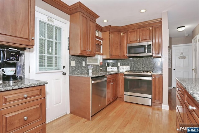 kitchen featuring stainless steel appliances, light wood finished floors, brown cabinetry, decorative backsplash, and stone counters