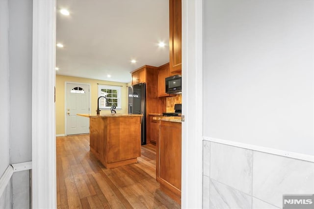 kitchen featuring black appliances, an island with sink, wood-type flooring, brown cabinetry, and recessed lighting
