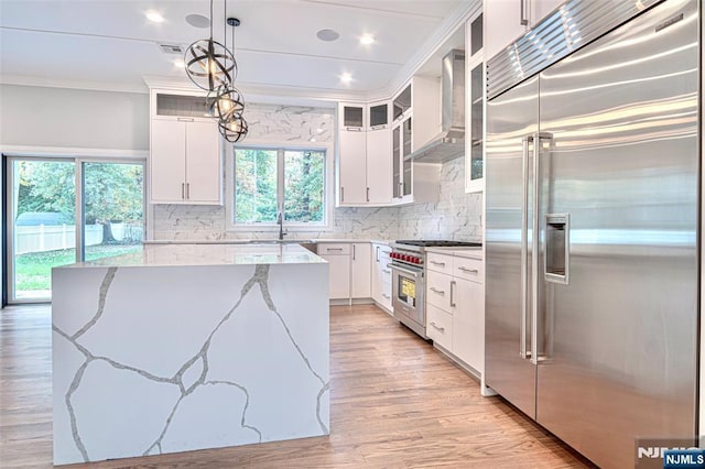 kitchen with premium appliances, wall chimney range hood, crown molding, light stone countertops, and white cabinets