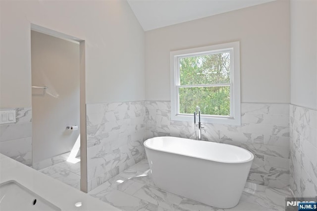 full bathroom featuring tile walls, a wainscoted wall, a soaking tub, marble finish flooring, and lofted ceiling