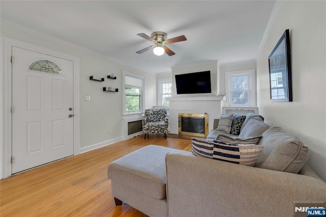 living area featuring ornamental molding, a brick fireplace, light wood finished floors, a ceiling fan, and baseboards