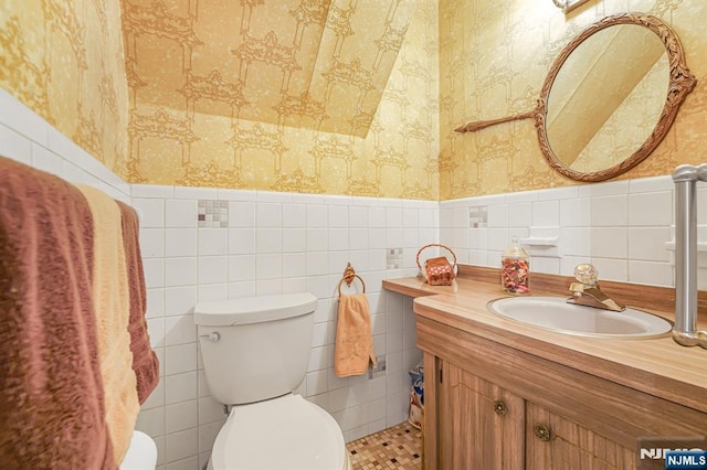 half bath with wallpapered walls, toilet, a wainscoted wall, vanity, and tile walls