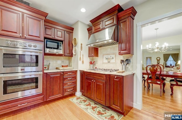 kitchen with under cabinet range hood, appliances with stainless steel finishes, light wood-type flooring, a chandelier, and tasteful backsplash