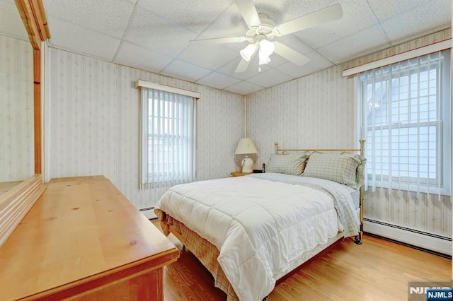 bedroom with a baseboard heating unit, a paneled ceiling, multiple windows, wood finished floors, and wallpapered walls