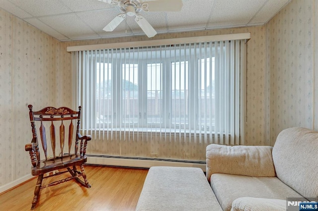 sitting room with wood finished floors, baseboard heating, wallpapered walls, ceiling fan, and a drop ceiling
