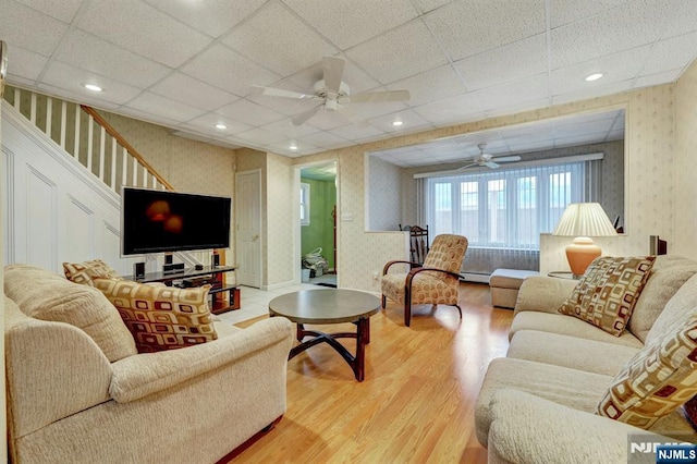 living room with wallpapered walls, ceiling fan, wood finished floors, stairs, and baseboard heating