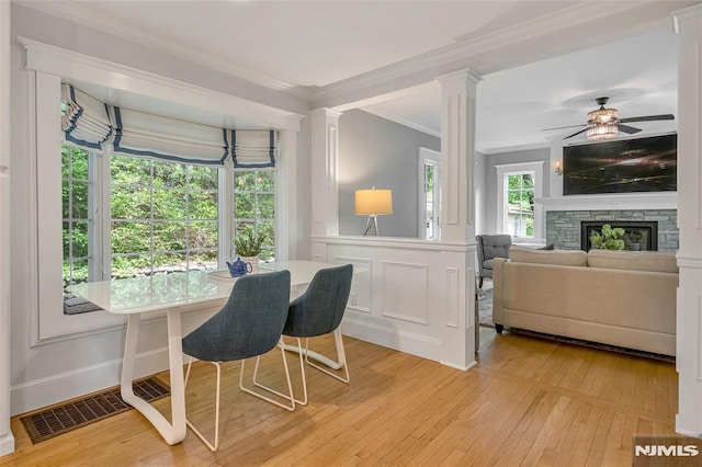 dining space featuring light wood-style flooring, crown molding, ornate columns, ceiling fan, and a stone fireplace
