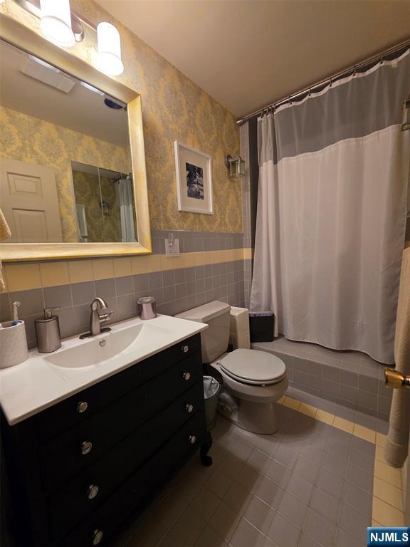 bathroom with wallpapered walls, tile patterned floors, toilet, vanity, and tile walls