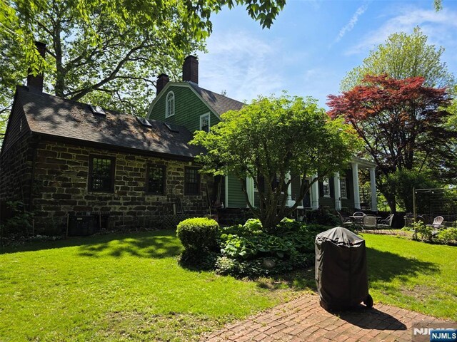 back of house featuring a lawn, stone siding, a chimney, a shingled roof, and a patio area